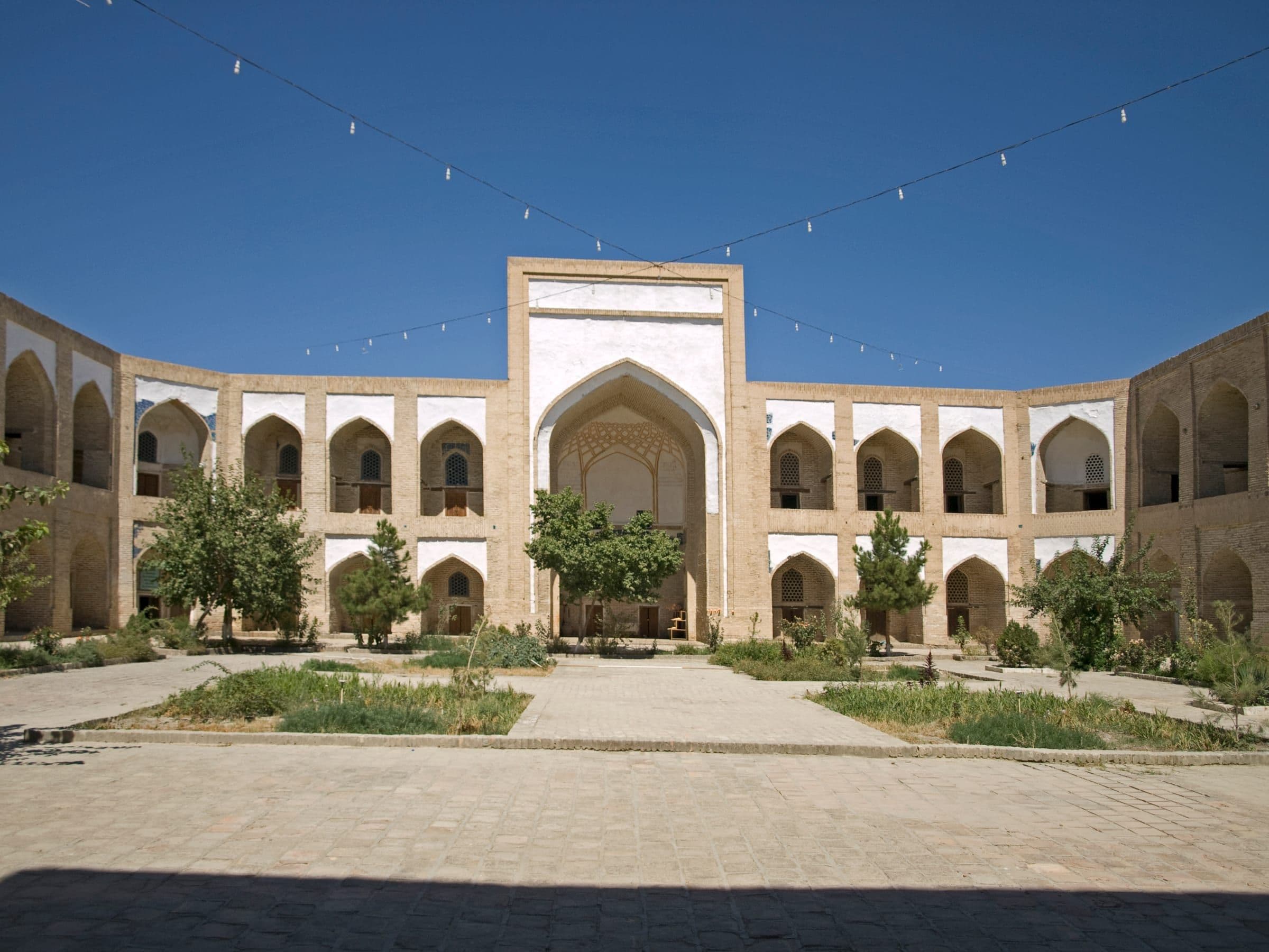 Arcaded courtyard in Bukhara: two storeys of pointed arches flanking a tall central portal, with young trees and paved paths