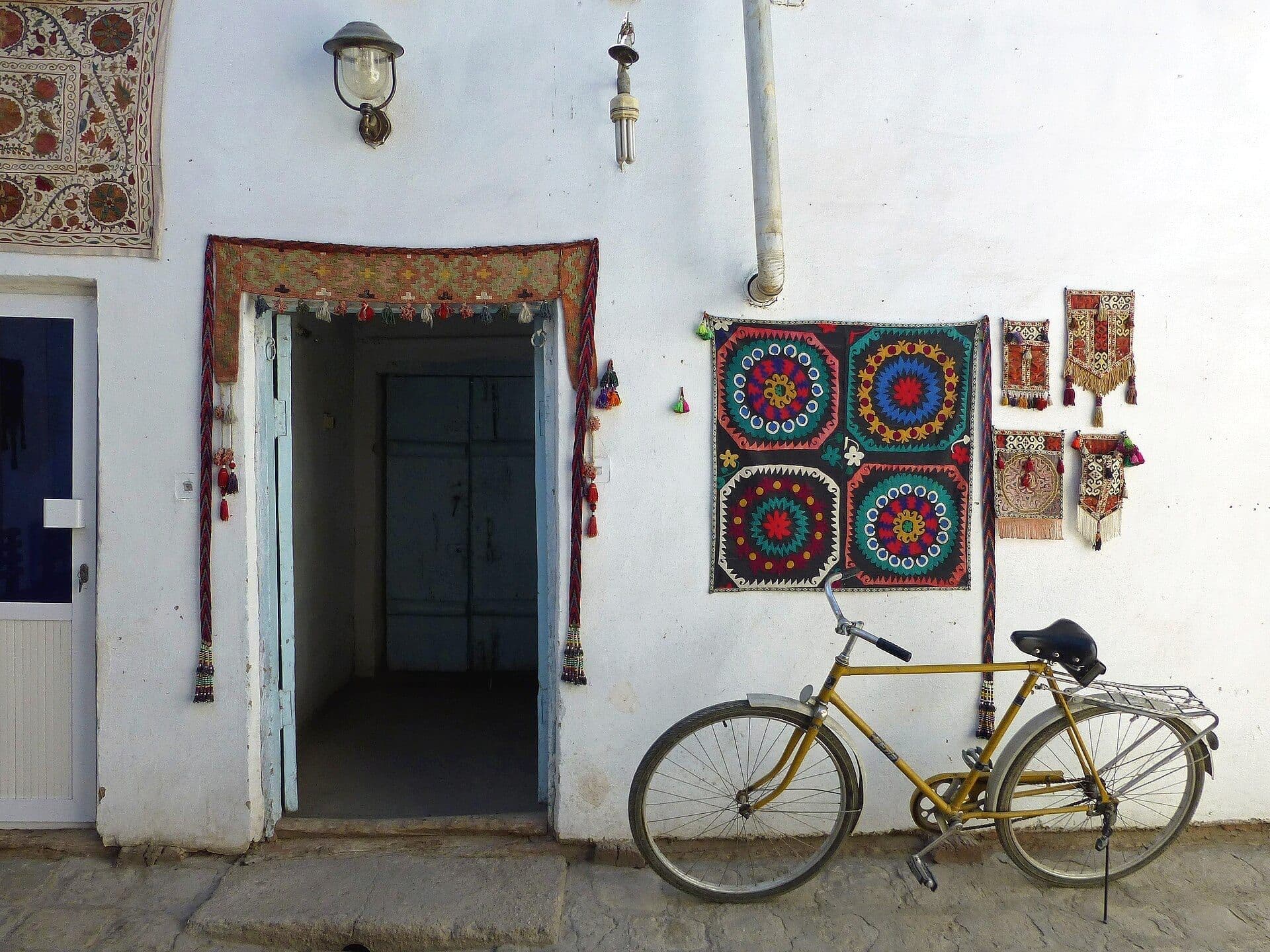 Bright suzani embroideries and tasselled hangings on a whitewashed wall by an open doorway, with an old yellow bicycle