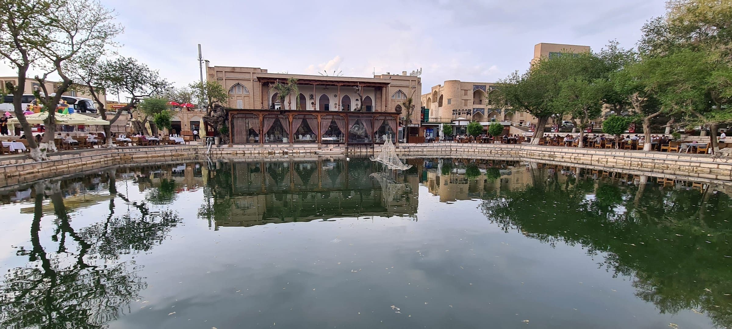 Buxorodagi Labi Hovuz: tosh qirg‘oqli hovuz bo‘yida daraxtlar va kafe stollari, suvda atrofdagi binolar aks etgan