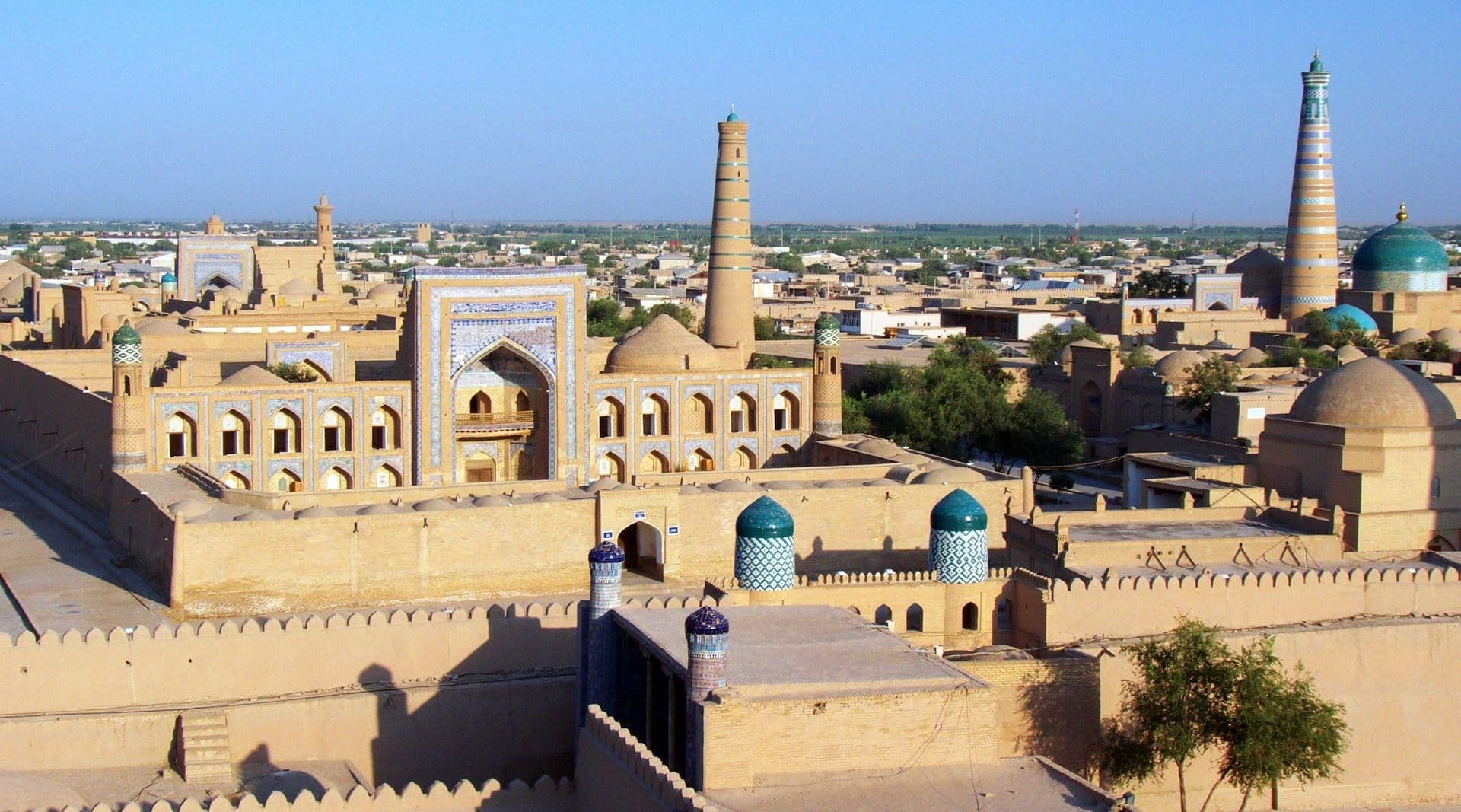 Devor bilan o‘ralgan eski Xiva tomlari: koshinli madrasa peshtoqi, gumbazlar va baland yo‘l-yo‘l minoralar