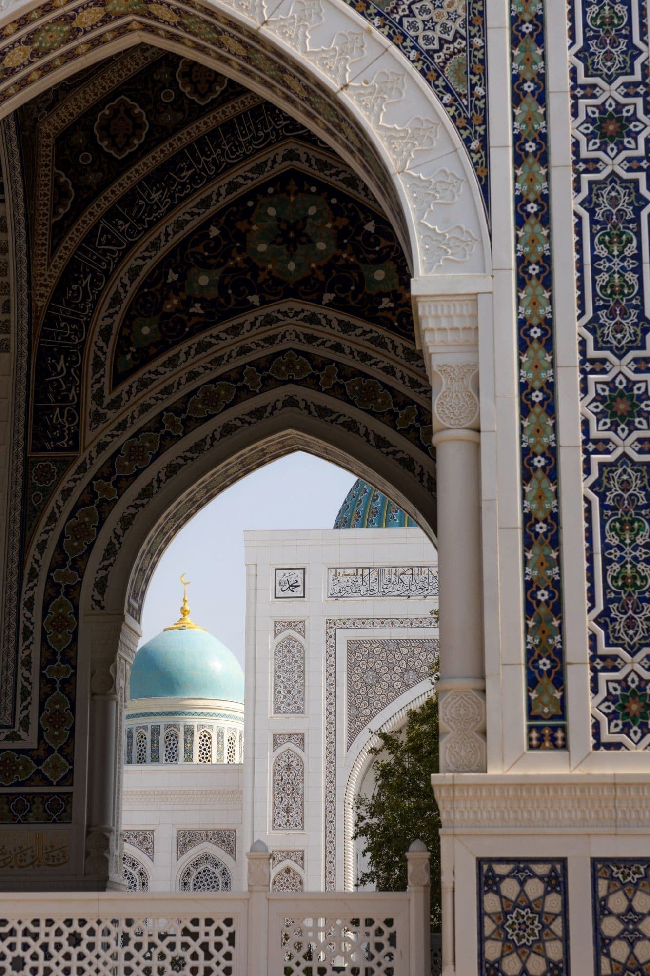 An ornate arch with calligraphy on its dark blue vault frames white stone walls and a turquoise dome with a gold crescent