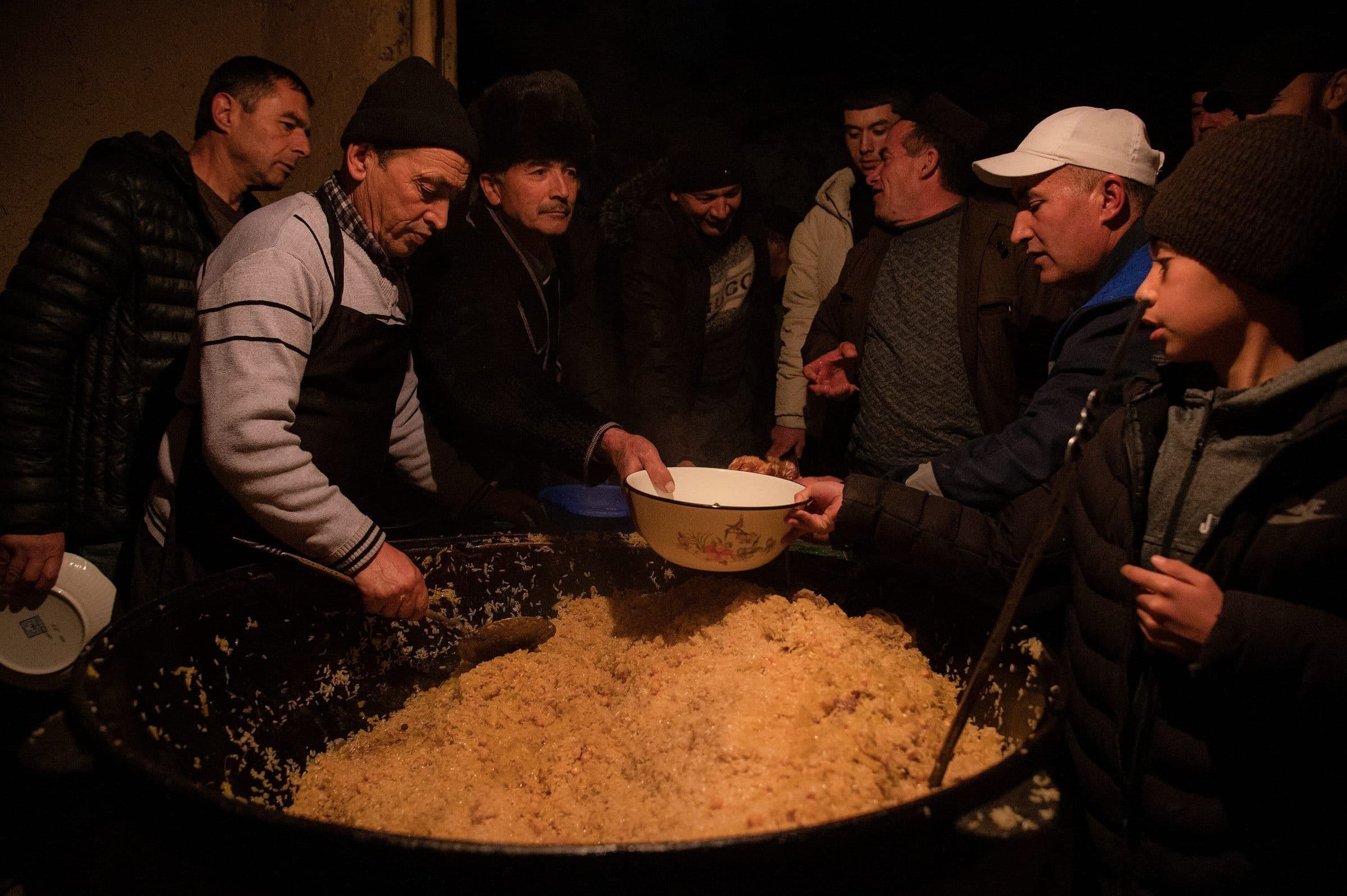 Men and a boy crowd around a huge kazan of plov as bowls are held out to the cook in an apron