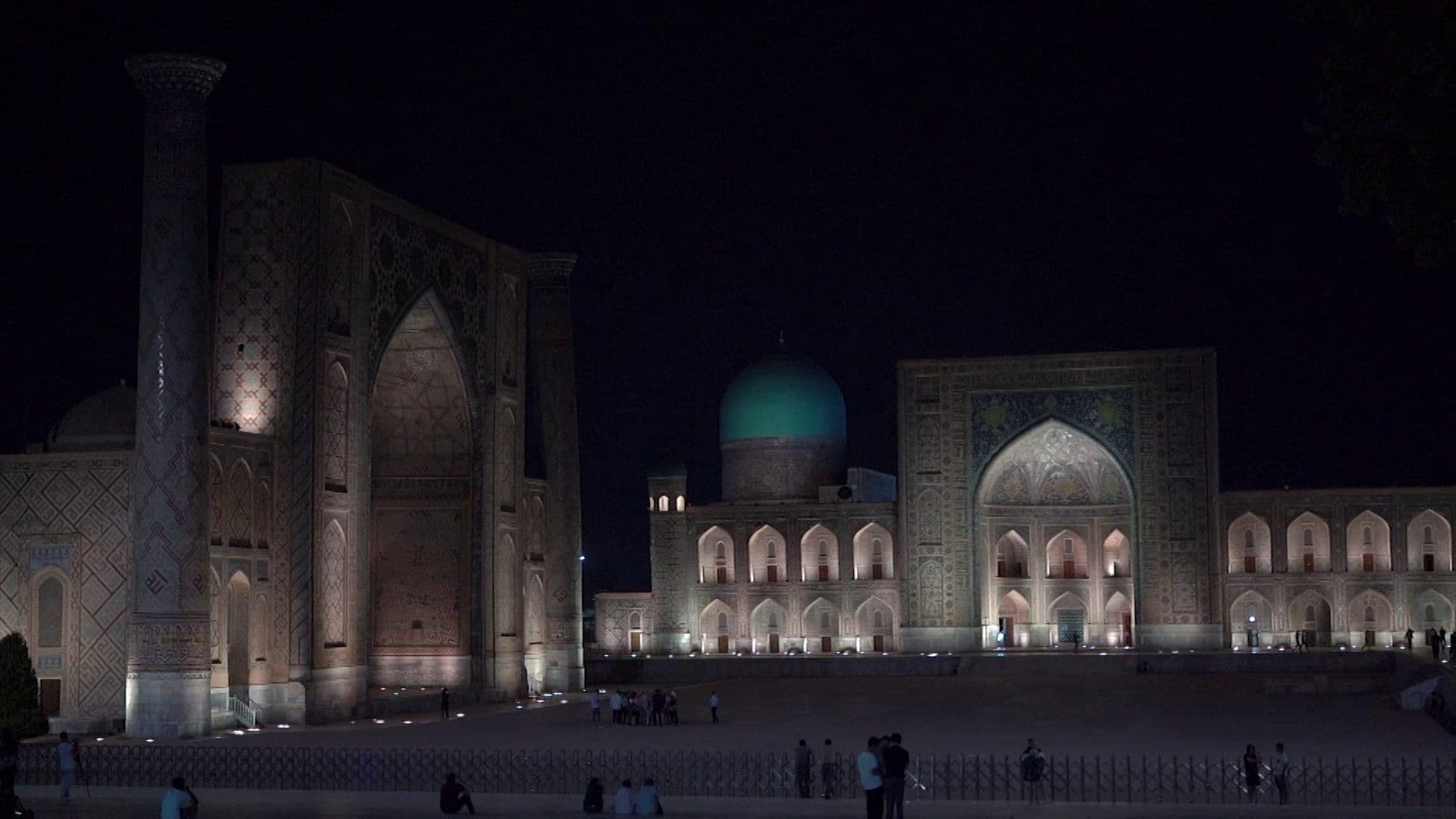 Registan, Samarkand, at night: floodlit madrasa portals and a turquoise dome above a dark square with a few visitors