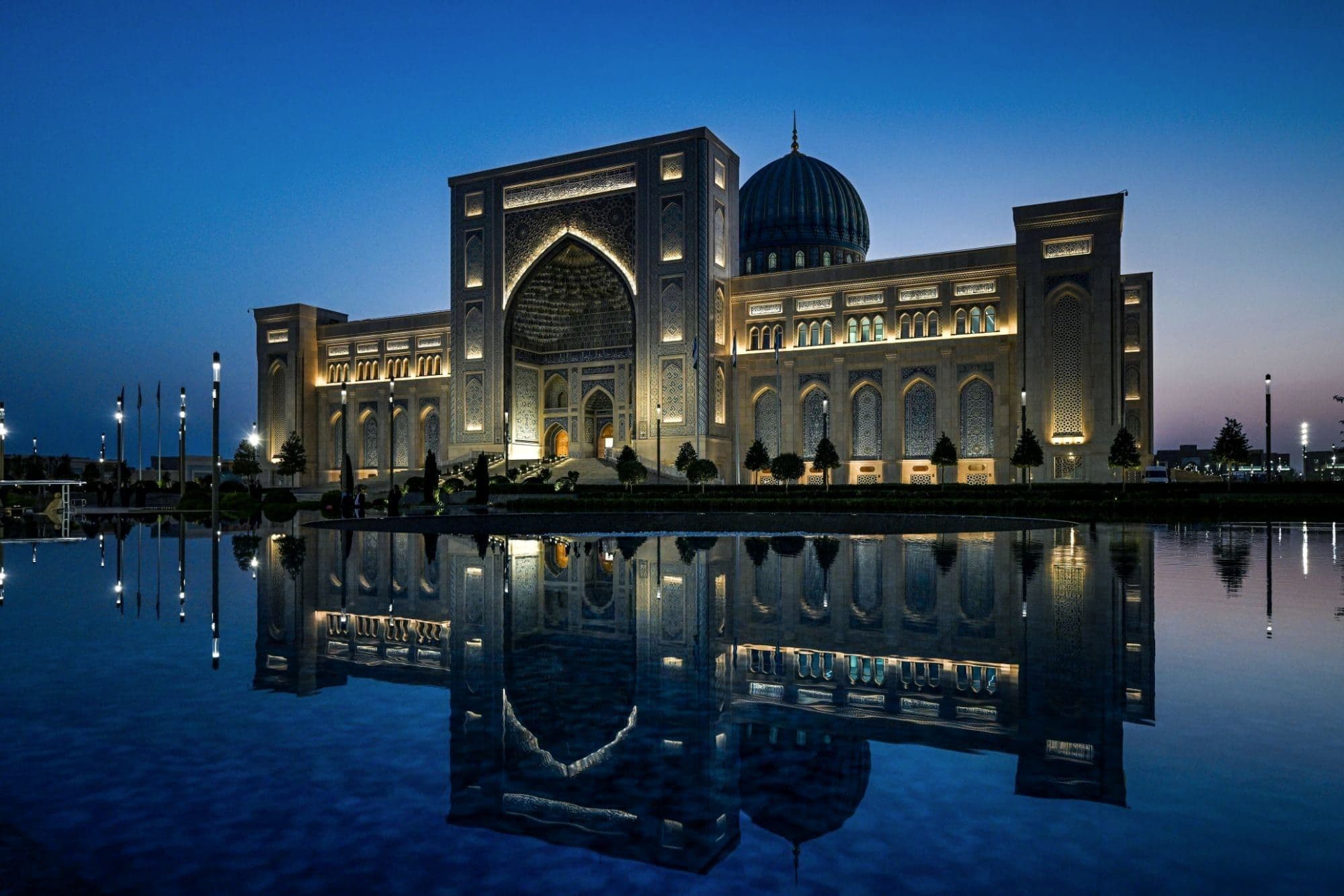 A grand building in Tashkent with a tall arched portal and blue dome, floodlit in deep blue twilight and mirrored in a pool